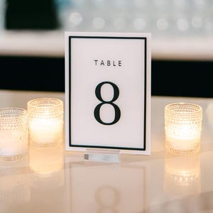 Puede incluir: Una tarjeta blanca con el número de mesa con un borde negro y el número 8. La palabra "TABLE" está encima del número. Tres velas votivas encendidas en portavelas de vidrio texturizado están frente a la tarjeta, creando un ambiente cálido.