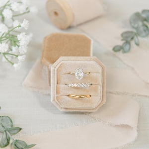 Op de afbeelding: Een beige fluwelen ringendoosje met drie gouden ringen: één met een ovaal geslepen middensteen, één met een rij kleine ronde steentjes en één gladde gouden ring. Het doosje is omringd door witte gipskruidbloemen en een beige zijden lint.