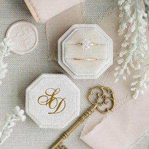 May include: Two white velvet ring boxes with gold lettering. One box has the initials "S&D" and the other has a diamond engagement ring and a thin gold band inside. A gold key and a spool of light pink ribbon are also in the image.