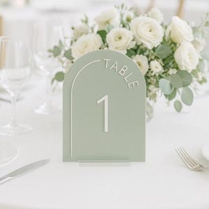 以下が含まれることがあります： 「TABLE」の文字と数字の「1」が白で書かれた、セージグリーンのテーブルナンバー。 テーブルナンバーは上部がアーチ型で、透明なベースの上に置かれています。 背景には白いバラのブーケがあります。