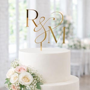 May include: A three-tiered white wedding cake with floral arrangements of pink and white roses and greenery. A gold-colored topper displays the letters "R & M". The cake is set in a bright, airy room, suggesting a celebration.