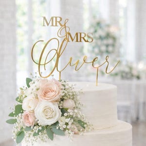 Peut inclure: Gâteau de mariage blanc à trois étages, orné de fleurs fraîches et d'un cake topper doré « Mr & Mrs Oliver ». Le gâteau est décoré de roses roses et blanches, de gypsophile et de verdure.