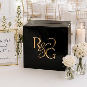 May include: A black square card box with gold R&G lettering, accompanied by a gold-framed sign reading CARDS and GIFTS THANK YOU, surrounded by white floral arrangements in glass vases and pillar candles in clear glass cylinders.