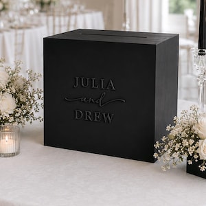 Könnte beinhalten: Eine quadratische, mattschwarze Kartenbox mit dem geprägten Schriftzug JULIA and DREW auf der Vorderseite. Die Box hat einen horizontalen Schlitz an der Oberseite und ist von weißen Blumenarrangements, drei schwarzen Stabkerzen und zwei Teelichtern umgeben.