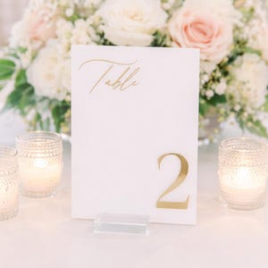 May include: A white table number card with gold lettering that reads "Table" and the number "2". The card is displayed on a clear acrylic stand, with lit candles in glass votives. A floral arrangement of white and pink roses is in the background.