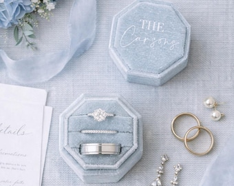 May include: Two octagonal light blue velvet ring boxes, one closed with the text THE Carsons, one open holding three rings. Includes two gold bands, two drop earrings with clear stones, white wedding stationery, and a blue floral arrangement.