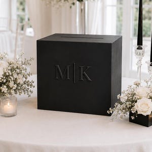 Op de afbeelding: Een matzwarte houten kaartenbox met een horizontale gleuf aan de bovenkant en de in reliëf aangebrachte letters M|K aan de voorzijde. De box is omringd door witte bloemstukken, glazen theelichthouders met brandende kaarsen en drie zwarte dinerkaarsen.