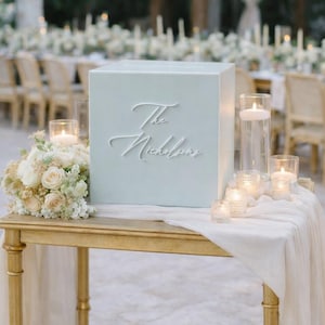 Puede incluir: Una caja de tarjetas cuadrada de color azul pálido con el texto en cursiva blanca The Nicholsons, sobre una mesa dorada con un camino de mesa blanco, un ramo de rosas blancas y varios portavelas de cristal con velas blancas encendidas.