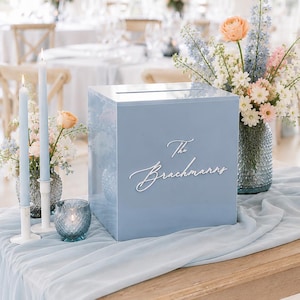 Peut inclure: Une boîte à cartes en acrylique bleu clair avec une fente sur le dessus et le texte en écriture blanche The Brachmanns sur le devant. La boîte est présentée avec des bougies effilées bleu clair, un photophore bleu et un bouquet dans un vase bleu.