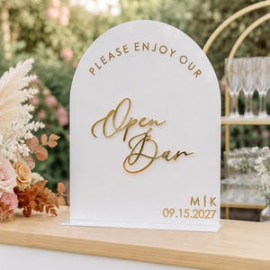 May include: A white arched sign with gold lettering that reads "PLEASE ENJOY OUR Open Bar" and "M|K 09.15.2027". The sign features a smooth surface and is positioned upright on a flat base, displaying a clean design with elegant, raised script text.