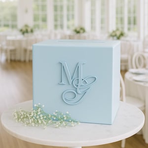 Op de afbeelding: Een lichtblauwe vierkante kaartenbox met een verhoogd monogram met de letters M en G op het voorpaneel. De doos heeft een horizontale gleuf aan de bovenzijde en is versierd met een klein takje witte gipskruidbloemen aan de onderkant.