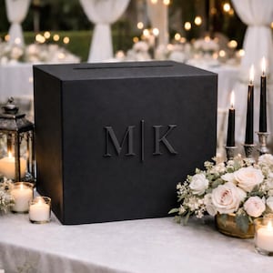 May include: A black wedding card box with the initials "MK" embossed on the front. The box sits on a white tablecloth, surrounded by floral arrangements, candles, and a lantern. The background features a blurred outdoor wedding setting.