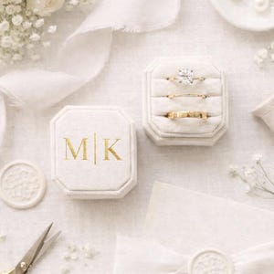 May include: An octagonal cream-coloured velvet ring box with the gold embossed letters M|K, containing three rings. The set includes a solitaire diamond ring, a thin band, and a wider gold band, displayed alongside gold scissors and a wax-sealed envelope.