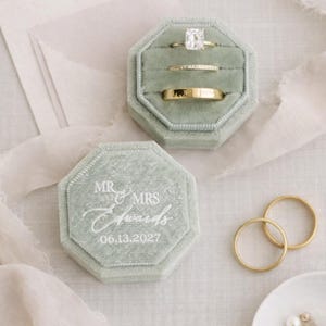 May include: Sage green velvet ring boxes with gold wedding rings. One box is open, displaying three rings: a diamond solitaire, a diamond band, and a gold band. The other box is closed, with the text "MR & MRS Edwards 06.13.2027".