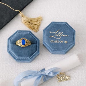 May include: A gold-colored class ring with a blue stone, displayed in a blue velvet octagonal box. The box is next to a matching box with the name "Lillian" and "CLASS OF '26" in gold lettering. A graduation cap tassel and a scroll with a "2026" charm are also present.