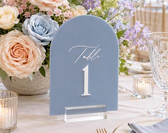 Könnte beinhalten: Ein staubblaues, bogenförmiges Acryl-Tischschild mit dem weißen Text Table 1, das auf einem klaren rechteckigen Sockel steht. Das Schild befindet sich neben einem Blumenarrangement aus pfirsichfarbenen und hellblauen Rosen und einem Glas.