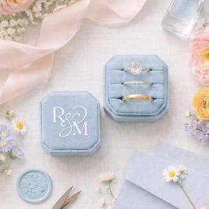May include: A light blue velvet ring box with the initials "R & M" in white, next to a box with three rings. The scene includes flowers, stationery, and gold scissors, creating a romantic, wedding-themed aesthetic.