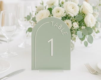 Könnte beinhalten: Ein salbeigrünes, bogenförmiges Tischnummernschild mit dem weißen Text TABLE und der Zahl 1. Das Schild hat eine weiße, bogenförmige Grafik und wird von einem transparenten Acrylsockel gehalten, der auf einer weißen Fläche steht.