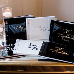 May include: Four black and white acrylic guest books with gold lettering. The books are on a table with candles and a gold surface. The books have the names of the couples and the date of their wedding.