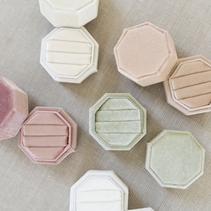 May include: A collection of octagonal velvet ring boxes in various pastel shades including cream, dusty rose, mauve, and sage green. Each box features a removable lid and a slotted interior designed to hold rings securely in place.