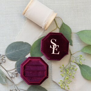 May include: An octagonal deep burgundy velvet ring box with the letters SE embossed in gold on the lid, accompanied by a wooden spool of frayed cream-colored ribbon and several green eucalyptus leaves with small clusters of buds.
