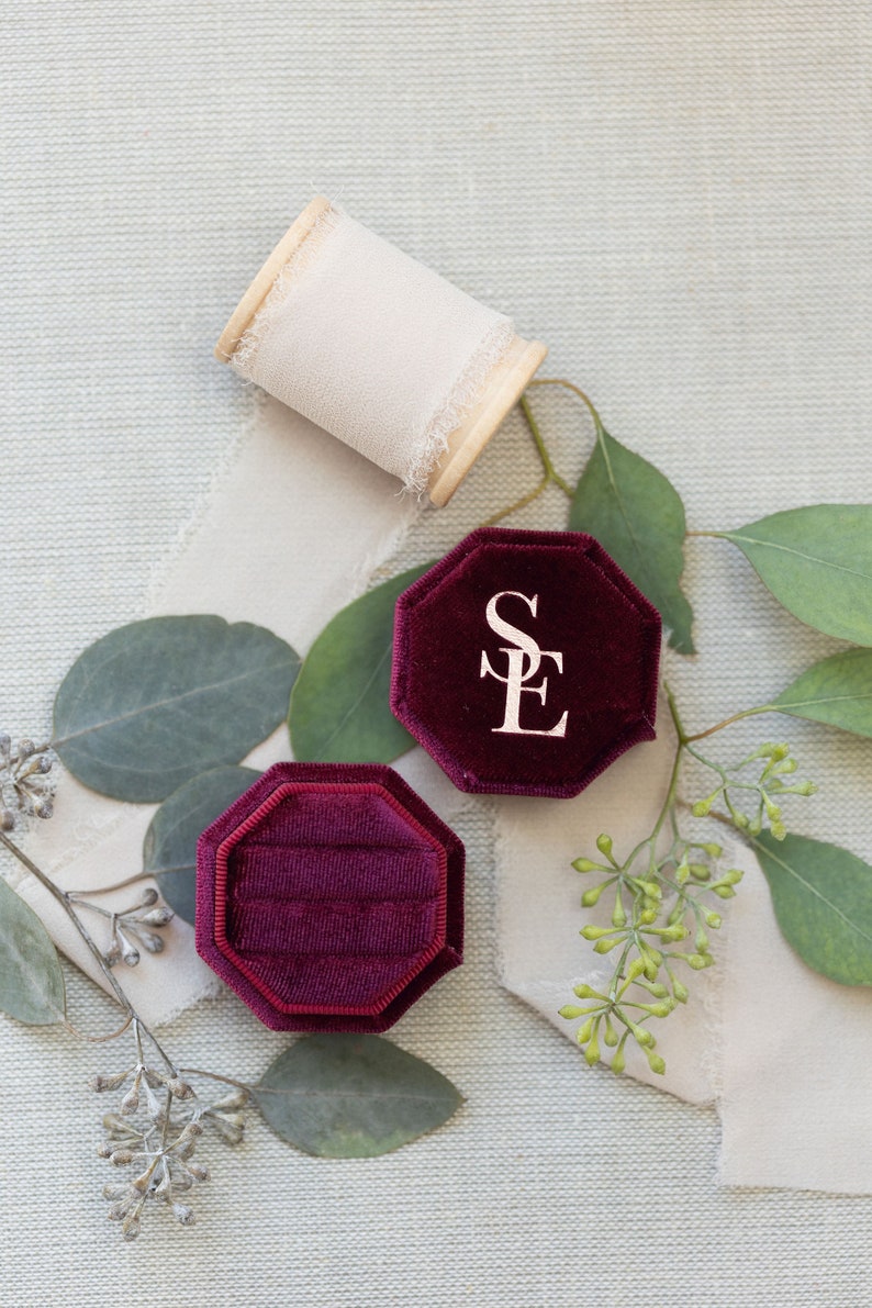 May include: An octagonal deep burgundy velvet ring box with the letters SE embossed in gold on the lid, accompanied by a wooden spool of frayed cream-colored ribbon and several green eucalyptus leaves with small clusters of buds.