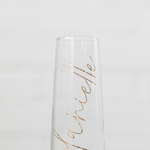May include: A clear glass champagne flute with the word "Brielle" written in gold lettering.