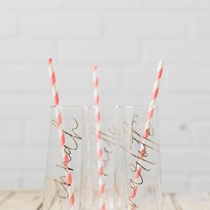 May include: Three clear glass champagne flutes with pink and white striped straws. Each flute has a gold inscription: "Krista", "Michelle", and "Angela Tottie".