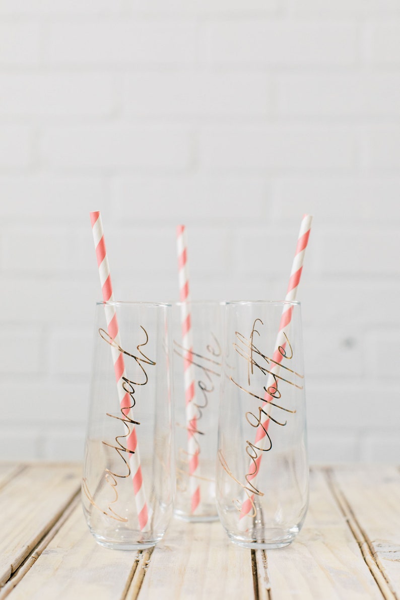 May include: Three clear glass champagne flutes with pink and white striped straws. Each flute has a gold inscription: "Krista", "Michelle", and "Angela Tottie".