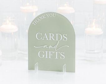 May include: An arched sage green acrylic sign featuring white text that reads THANK YOU CARDS and GIFTS. The sign is supported by two clear acrylic stands, providing a stable base for the display of this event signage piece.