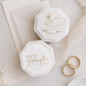 May include: An octagonal, light gray ring box with the inscription "MR & MRS. Johnston 11.5.2027" in gold lettering. The box is open, revealing three rings. Two gold wedding bands and a small dish with pearl earrings are also visible.