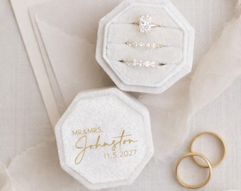 May include: An octagonal white velvet ring box with gold text reading MR&MRS. Johnston 11.5.2027. The open box holds three rings, including one with a large rectangular stone and two bands with small stones. Two plain gold bands sit nearby.