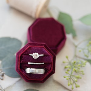 May include: A deep burgundy velvet octagonal ring box containing three rings: a solitaire diamond ring with a pav&eacute; band, a thin diamond eternity band, and a wide, brushed silver-toned metal band, all nestled in the plush fabric interior.