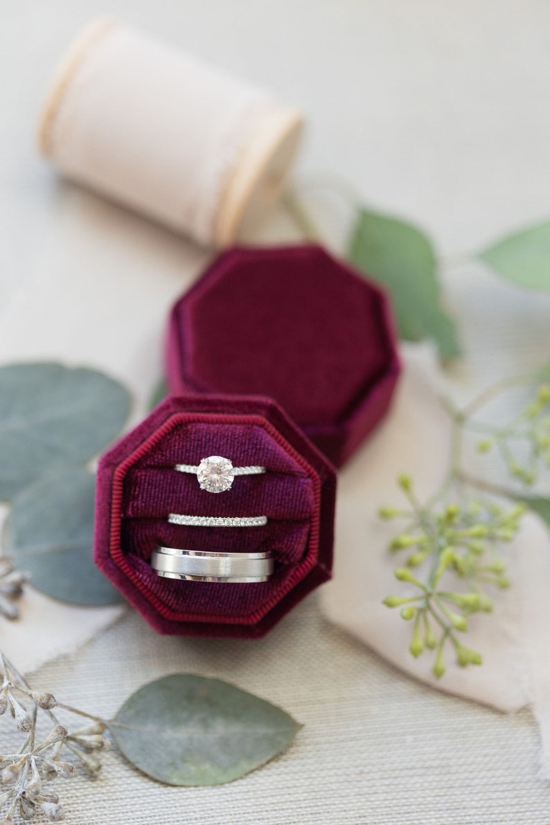 May include: A deep burgundy velvet octagonal ring box containing three rings: a solitaire diamond ring with a pav&eacute; band, a thin diamond eternity band, and a wide, brushed silver-toned metal band, all nestled in the plush fabric interior.