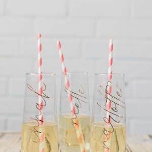 May include: Three clear glass champagne flutes with pink and white striped straws. Each flute has a gold inscription: "Sarah", "Michelle", and "Ashley".