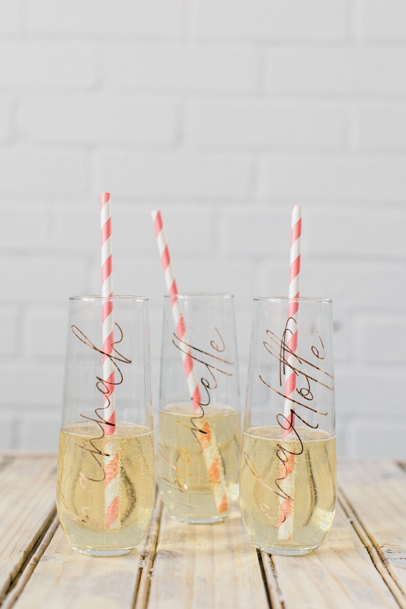 May include: Three clear glass champagne flutes with pink and white striped straws. Each flute has a gold inscription: "Sarah", "Michelle", and "Ashley".
