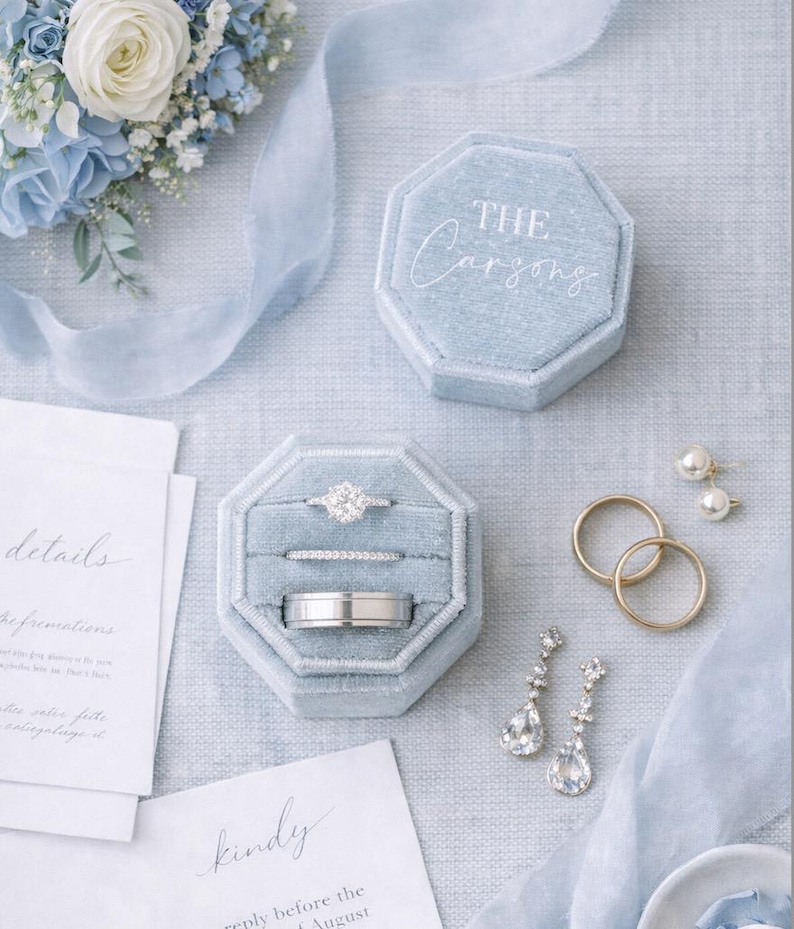 May include: Two octagonal light blue velvet ring boxes, one closed with the text THE Carsons, one open holding three rings. Includes two gold bands, two drop earrings with clear stones, white wedding stationery, and a blue floral arrangement.