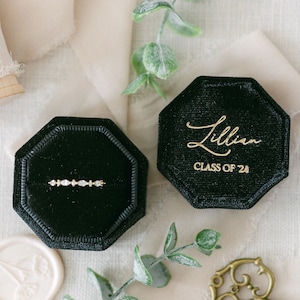 Könnte beinhalten: Ein goldener Ring mit einer Reihe von klaren Steinen im Marquise- und Rundschliff, platziert in einer achteckigen schwarzen Samtschachtel. Der Deckel trägt die goldene Aufschrift Lillian CLASS OF '24. Ring und Schachtel sind mit Grünzeug dekoriert.