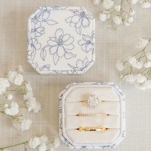 May include: An octagonal ring box with a white and blue floral pattern containing three gold rings, including one with an oval-cut stone and one with small pavé diamonds, resting on a cream-colored fabric insert.