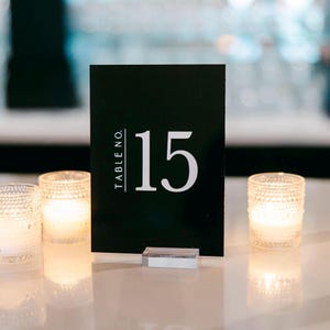 Op de afbeelding: Zwarte tafelnummerkaart met witte tekst die "TABLE NO. 15" aangeeft.
