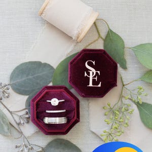 May include: A deep burgundy velvet octagonal ring box with the letters SE embossed in white on the lid. The open box contains three silver-toned rings, including one with a central stone and two bands. A spool of cream ribbon is nearby.