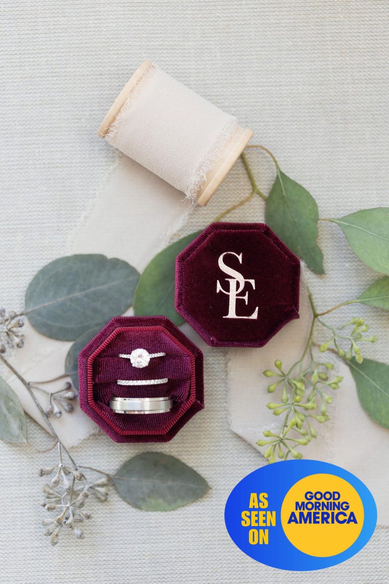 May include: A deep burgundy velvet octagonal ring box with the letters SE embossed in white on the lid. The open box contains three silver-toned rings, including one with a central stone and two bands. A spool of cream ribbon is nearby.