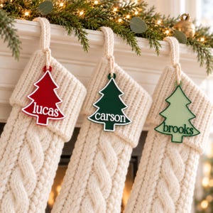 May include: Three cream-colored cable knit stockings hanging with personalized tree-shaped ornaments. The red ornament reads lucas, the dark green ornament reads carson, and the light green ornament reads brooks in white lettering.