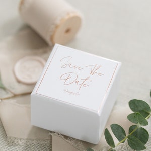 May include: A square white box with a rose gold border and the text Save The Date printed in rose gold script. The box is accompanied by a spool of beige frayed ribbon, a circular wax seal, and a sprig of green eucalyptus leaves.