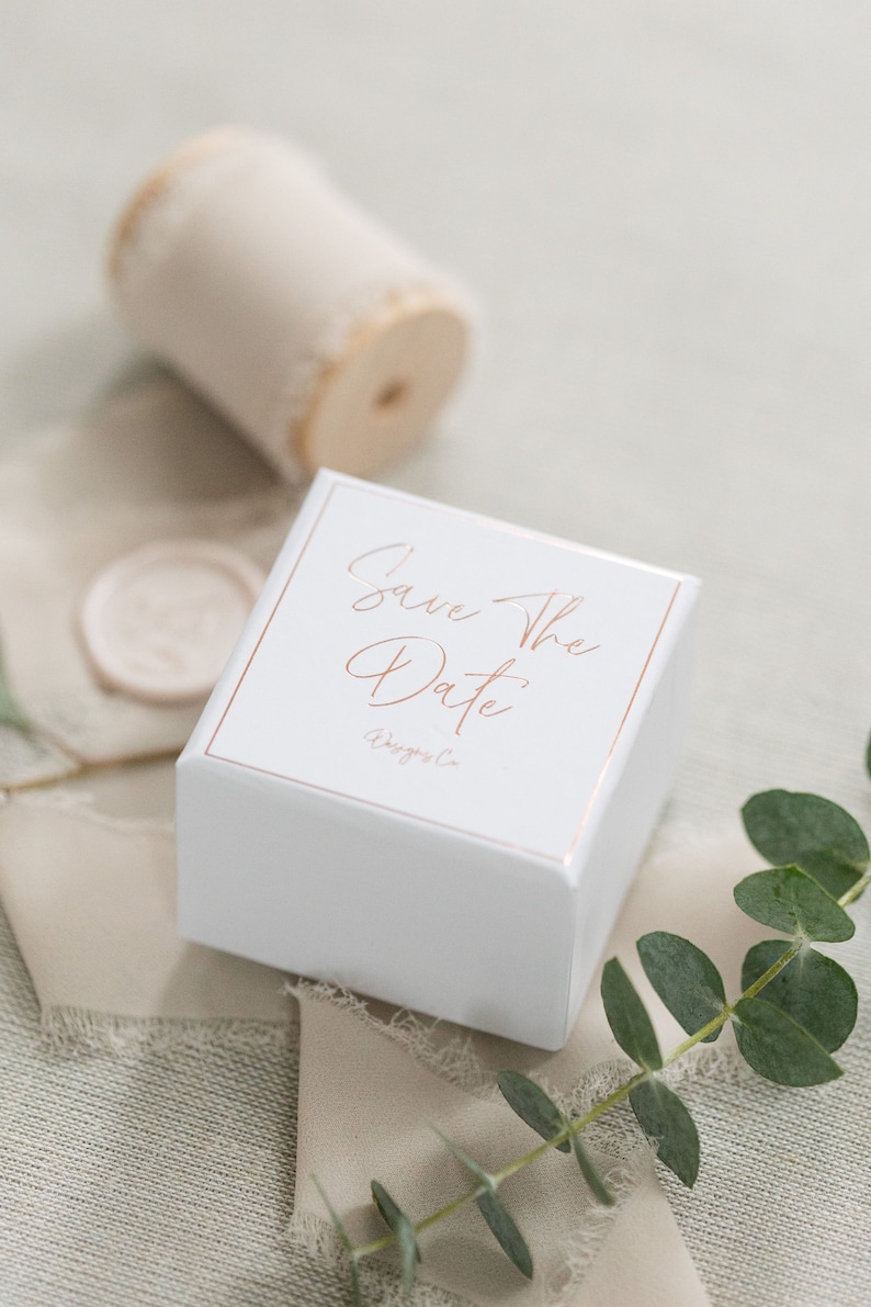 May include: A square white box with a rose gold border and the text Save The Date printed in rose gold script. The box is accompanied by a spool of beige frayed ribbon, a circular wax seal, and a sprig of green eucalyptus leaves.