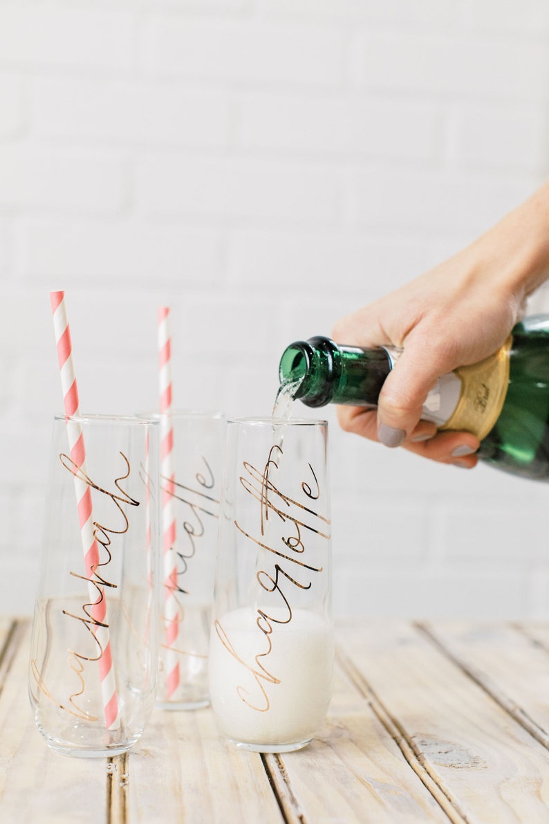 May include: Three champagne flutes with pink and white striped straws. The flutes are filled with champagne and have gold lettering that reads "Kathryn", "Michelle", and "Charlotte".