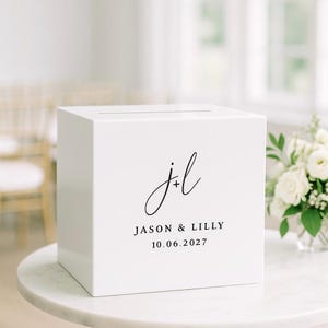 Op de afbeelding: Een witte vierkante kaartenbox met een horizontale gleuf aan de bovenkant. De voorzijde is voorzien van zwarte tekst met de initialen J+L in een cursief lettertype, gevolgd door de namen JASON & LILLY en de datum 10.06.2027 in een strak lettertype.