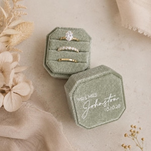 Pode incluir: Uma caixa de anéis octogonal em veludo verde sálvia com três ranhuras contendo três anéis de ouro. A tampa apresenta o texto impresso em branco MR & MRS Johnston 10.10.28. A ranhura superior contém um anel solitário oval, a do meio uma aliança de diamantes e a inferior uma aliança de ouro simples.