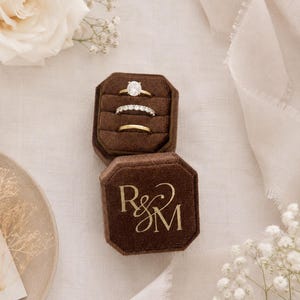 May include: An octagonal dark brown velvet ring box with the gold monogram R&M printed on the lid. The open box contains three rings: a gold ring with a large oval stone, a silver-toned band with small round stones, and a plain gold band.