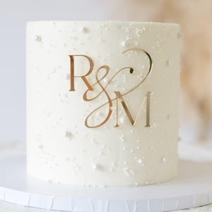 Puede incluir: Un pastel cilíndrico blanco con un acabado glaseado suave, decorado con perlas blancas dispersas y un adorno central de acrílico dorado para pasteles que muestra las letras estilizadas R y M unidas por un símbolo ampersand.